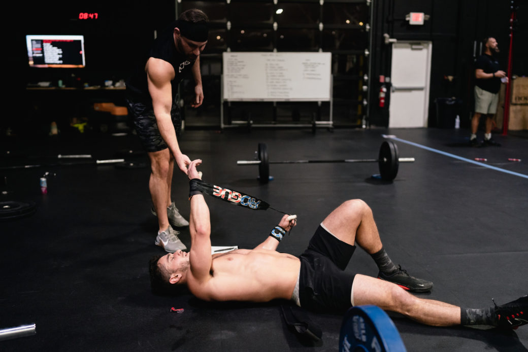 A man fist-bumps another man lying on the floor after a workout. 